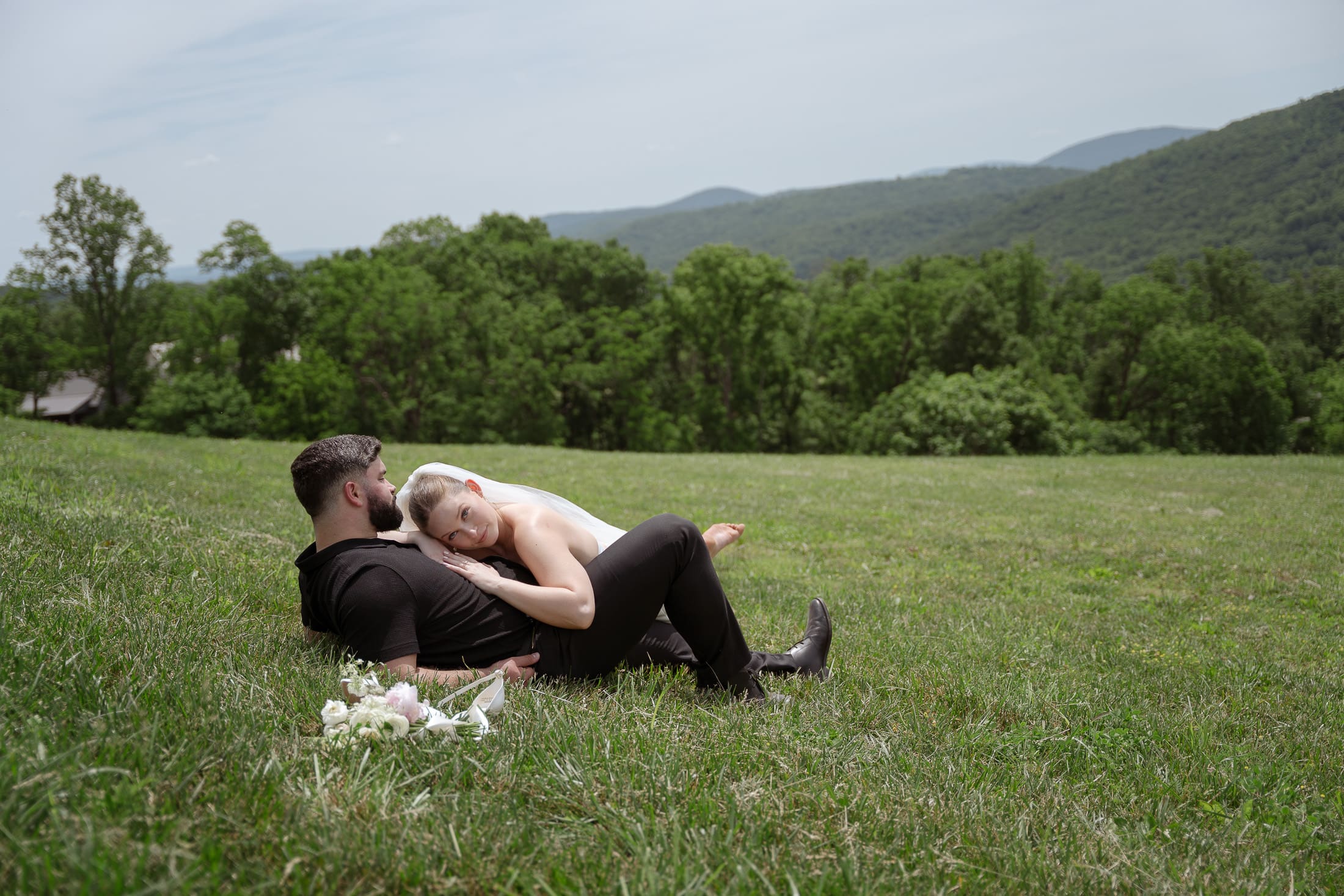 Bride lays on groom's chest as they lay in a large grass field during their hiking elopement day.